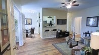This single-floor residence offers a thoughtfully designed open-concept layout that connects the living, dining, and kitchen areas into one cohesive and functional living space. Warm hardwood flooring flows throughout, lending a sense of continuity and natural warmth to the interior.

The living area is anchored by a ceiling fan with integrated lighting, providing both comfort and illumination. A wall-mounted flat-screen television and a low-profile media console serve as focal points, while a glass-top coffee table and area rug define the seating zone.

A peninsula-style breakfast bar with dark granite countertops and sleek bar stools separates the kitchen from the main living space. The kitchen is visible through a pass-through window, revealing white cabinetry, stainless steel appliances including a microwave and refrigerator, and ample counter space — ideal for everyday cooking and entertaining.

Adjacent to the kitchen, the dining area accommodates a full dining table and chairs, framed by a decorative wall mirror and a charming lattice-panel room divider that adds architectural character. Natural light filters through the space, enhancing the bright and airy ambiance. The layout is efficiently organized, with interior doors leading to additional private areas of the residence.