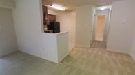 This single-floor residential unit offers a functional and straightforward layout designed for comfortable everyday living. The kitchen is positioned at the heart of the space and features a raised pass-through counter that opens toward the living area, promoting an open and connected feel. The kitchen is equipped with dark wood cabinetry, a black refrigerator, and a stovetop, along with overhead fluorescent lighting that keeps the workspace well-lit. A fire extinguisher is visibly mounted, reflecting attention to safety. The adjoining living area is finished with wall-to-wall carpeting in a neutral beige tone, contributing to a warm and quiet atmosphere. The light-colored walls throughout create a clean, understated backdrop that is easy to personalize. A hallway extends from the living space, leading to additional rooms, including what appears to be a bathroom with a visible tub and closet space. The layout flows efficiently from one area to the next, making practical use of the available square footage. Ceiling-mounted lighting fixtures provide consistent illumination throughout. Overall, the unit presents a no-frills, move-in-ready living environment suited for those seeking a clean and functional single-level home.