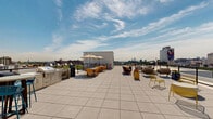 Located at 1201 Callowhill St in Philadelphia's vibrant Callowhill neighborhood, this rooftop deck offers a dynamic and well-appointed outdoor amenity space with striking panoramic views of the city skyline. The layout is divided into distinct functional zones, seamlessly blending relaxation and social gathering areas across a generously sized open-air terrace.

A standout feature is the lush artificial turf section, dotted with modern chaise lounges and side tables, providing a comfortable setting for sunbathing while taking in sweeping views of Philadelphia's skyline, including recognizable landmarks and colorful local murals. The turf area is bordered by sleek metal railings that maintain open sightlines to the surrounding cityscape in all directions.

Adjacent to the turf zone is a hard-surface terrace area furnished with modular seating arrangements, including sofas and lounge chairs, ideal for casual gatherings. A covered indoor-access structure anchors the rooftop, connecting the space to the building below. Further along the deck, a shaded dining and lounge area features market umbrellas and colorful seating, offering a more protected setting for outdoor entertaining.

Planters with seasonal flowers add touches of color throughout, while the unobstructed 360-degree views of Philadelphia's urban fabric — from historic brick buildings to modern towers — make this rooftop a compelling and distinctive outdoor destination.