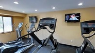 This single-level fitness center is designed to provide a straightforward and functional exercise environment. The space features a selection of cardio equipment, including treadmills and elliptical trainers from Life Fitness, a well-regarded commercial brand known for its durable and user-friendly machines. Each piece of equipment is equipped with its own digital display console, allowing users to monitor workout metrics such as speed, incline, and elapsed time.

Two flat-screen televisions are mounted on the walls at viewing height, positioned to be visible from the cardio machines, enabling users to watch programming during their workouts. The walls are painted in a neutral warm beige tone, contributing to a clean and understated aesthetic. Recessed lighting in the white ceiling provides consistent, even illumination throughout the space.

The flooring appears to be a dark, rubberized surface, which is standard in fitness environments for its durability and shock-absorbing properties. The overall layout is efficient, making good use of the available floor space to accommodate multiple users simultaneously. The room presents a no-frills, practical setting suited for cardiovascular training.