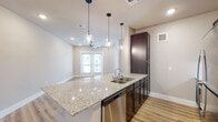 This single-floor residence showcases a thoughtfully designed open-concept layout that seamlessly connects the kitchen and living areas. The kitchen is a standout feature, centered around a generous granite-topped island with a double basin sink, stainless steel dishwasher, and a full-size stainless steel refrigerator. Rich dark-toned cabinetry contrasts beautifully against the light granite surfaces, while a herringbone-patterned tile backsplash adds a refined decorative touch. Three glass pendant lights hang above the island, providing focused task lighting and an elegant aesthetic detail.

The living area extends naturally from the kitchen, bathed in natural light from a set of French doors that open to the exterior. Recessed lighting and a ceiling fan overhead contribute to a comfortable, well-lit environment. Light-toned hardwood-style flooring runs consistently throughout the space, reinforcing the open, airy feel.

A bedroom features a window with blinds that allows natural light to filter in, while maintaining privacy. The bathroom is well-appointed with a granite-top vanity featuring dark cabinetry, a subway-tiled tub-shower combination, and wood-style flooring. Neutral warm-beige walls throughout the unit create a cohesive, clean, and modern aesthetic.