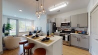 Located at 555 Dresher Rd in Horsham, Pennsylvania, this single-floor residence offers a thoughtfully designed open-concept layout with modern finishes throughout. The kitchen serves as a central hub of the living space, featuring grey cabinetry with sleek hardware, a subway tile backsplash, stainless steel appliances including a gas range, microwave, and refrigerator, and a large island with quartz countertops, an undermount sink, and pendant globe lighting overhead. Light hardwood flooring flows seamlessly from the kitchen into the open living area, which benefits from recessed lighting and large windows that invite natural light and outdoor views. The living room provides a comfortable, well-lit retreat with access to a balcony. Two bedrooms are carpeted and outfitted with oversized windows fitted with horizontal blinds, offering greenery views. The primary bedroom features a striking sputnik-style chandelier, while the secondary bedroom is equally bright and welcoming. The bathroom is finished with large-format marble-look tile flooring, a glass-enclosed walk-in shower with full tile surround, and a walk-in closet directly accessible from the bathroom. The overall ambiance is clean, contemporary, and well-appointed, reflecting a modern aesthetic with warm, neutral tones throughout.