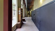 This parcel locker area is housed within a building of distinctive industrial character, where exposed ductwork, wooden-slat ceilings, and polished concrete floors create a raw yet refined aesthetic. The space is accessed through a light-filled entry vestibule framed by floor-to-ceiling glass walls and aluminum-framed doors, offering views of an exterior courtyard defined by warm exposed brick walls and open-air walkways.

The main corridor is generously proportioned, with soaring ceilings that amplify the sense of openness. Large windows line one side of the hall, drawing in abundant natural light that illuminates the earthy tones of the concrete flooring. A bank of sleek, dark charcoal parcel lockers runs along one wall, providing secure and organized package storage in a variety of compartment sizes to accommodate deliveries of all scales.

Adding a welcoming community touch, a freestanding wooden bookshelf stocked with a curated collection of books stands alongside simple wooden bench seating — offering residents a casual browsing spot. Subtle wall-mounted lighting complements the natural light throughout the corridor, while safety equipment and signage are thoughtfully integrated without disrupting the overall aesthetic. The space balances practical utility with an inviting, loft-inspired atmosphere.