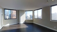 This single-floor space is situated at an elevated position within a high-rise building, offering sweeping views of a dense urban skyline. The main living area is defined by a series of large black-framed windows that wrap around a curved exterior wall, flooding the interior with abundant natural light throughout the day. The windows frame prominent city landmarks and neighboring high-rise buildings, providing a dynamic and ever-present urban backdrop.

The flooring throughout consists of dark hardwood, which contrasts cleanly with the crisp white ceilings and neutral greige-toned walls. The combination creates a contemporary and polished aesthetic. Clean white baseboards line the perimeter of the room, adding a refined finishing detail.

The open layout of the main area allows for flexible furnishing arrangements, with the angular geometry of the windowed wall adding architectural interest. The smooth, flat ceilings contribute to a clean, modern look, while the generous window placement ensures the space remains bright and visually connected to the cityscape outside. Overall, the space conveys a sleek, metropolitan character well-suited to an urban lifestyle.