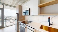 This thoughtfully designed one-bedroom, one-bathroom unit offers a compelling blend of industrial aesthetics and contemporary finishes across a compact, efficiently laid-out floor plan. The open-concept kitchen and living room is the heart of the space, anchored by floor-to-ceiling windows that frame expansive city skyline views and flood the area with natural light. The kitchen features a striking marble-veined quartz backsplash and countertops, light wood and white gloss cabinetry, stainless steel appliances including a range and refrigerator, and an integrated microwave — all set beneath a raw exposed concrete ceiling with visible ductwork that lends an industrial character throughout. Light hardwood flooring runs seamlessly through the living areas and bedroom, complementing the neutral-toned walls. The bedroom accommodates comfortable furnishings and benefits from a window with natural light, exposed concrete columns and ceiling, and access to two closets with built-in shelving. The combined bathroom and laundry room offers tiled flooring, a stacked washer/dryer unit, and practical fixtures. A hallway connects all areas of the unit efficiently. This space delivers a modern urban living experience with standout architectural details and striking high-floor views.
