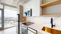 This thoughtfully designed one-bedroom, one-bathroom unit offers a compelling blend of industrial aesthetics and contemporary finishes across a compact, efficiently laid-out floor plan. The open-concept kitchen and living room is the heart of the space, anchored by floor-to-ceiling windows that frame expansive city skyline views and flood the area with natural light. The kitchen features a striking marble-veined quartz backsplash and countertops, light wood and white gloss cabinetry, stainless steel appliances including a range and refrigerator, and an integrated microwave — all set beneath a raw exposed concrete ceiling with visible ductwork that lends an industrial character throughout. Light hardwood flooring runs seamlessly through the living areas and bedroom, complementing the neutral-toned walls. The bedroom accommodates comfortable furnishings and benefits from a window with natural light, exposed concrete columns and ceiling, and access to two closets with built-in shelving. The combined bathroom and laundry room offers tiled flooring, a stacked washer/dryer unit, and practical fixtures. A hallway connects all areas of the unit efficiently. This space delivers a modern urban living experience with standout architectural details and striking high-floor views.