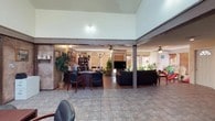 This single-floor space offers a thoughtfully arranged layout that blends professional functionality with a welcoming, lived-in atmosphere. The interior is defined by a mix of tile and hardwood-style flooring throughout, with textured, faux-painted walls in warm earth tones adding character and warmth to the environment.

The main area is expansive and open, divided into distinct zones by structural columns with exposed wood beam accents. A dedicated office section features multiple workstations with dark wood desks, ergonomic seating, shelving units, and computer setups, providing ample workspace for staff. Ceiling fans are installed throughout, contributing to airflow and comfort.

A comfortable lounge area is furnished with plush dark sofas, a decorative coffee table, and a flat-screen television, offering a relaxed setting adjacent to the workspace. Large windows dressed with wooden blinds allow natural light to filter through, brightening the interior considerably.

The kitchen area includes a bar-height counter with leather-upholstered barstools, dark upper cabinetry, and appliances including a coffee station and microwave. The textured ceiling and recessed lighting complete the functional yet inviting character of this well-appointed space.