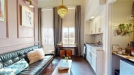 Located at 126 E Main Plaza in the heart of downtown San Antonio, this one-bedroom, one-bathroom unit blends historic charm with thoughtfully curated design. The living area immediately impresses with dramatically tall ceilings, floor-to-ceiling gray drape panels, and a stunning tiered gold crystal chandelier that serves as the room's focal centerpiece. Dark hardwood floors run throughout, grounding the space with warmth and character, while large windows flood the interior with natural light and offer views of the surrounding historic architecture.

The kitchen runs efficiently along one wall, featuring crisp white cabinetry with brushed gold hardware, a mosaic tile backsplash, granite-style countertops, and a stainless steel sink. A dedicated closet area provides additional storage and appliance space.

The bedroom is a serene retreat accented by rich terracotta-orange wall paneling, vibrant artwork, and generous windows that continue the theme of natural light. Two closets serve the space practically.

The bathroom is a charming highlight, finished with patterned teal-and-white geometric floor tiles, white subway wall tile, a white vanity with gold hardware, a round mirror with a brass frame, and a full soaking tub with shower. Every room reflects a cohesive aesthetic — eclectic, warm, and full of personality befitting its prime downtown San Antonio location.
