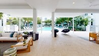 This expansive outdoor amenity area is designed with a seamless blend of covered and open-air spaces, centered around a large, crystal-clear swimming pool. The pool deck is lined with lounge chairs and teal umbrellas, framed by mature palm trees and tropical greenery that reinforce a resort-like atmosphere. Crisp white architecture and light travertine tile flooring unify the entire space, lending a clean, contemporary aesthetic throughout.

A generously sized covered pavilion extends from the pool area, supported by sleek white columns that define the space while maintaining an open, airy feel. This sheltered zone is thoughtfully divided into distinct areas for relaxing and socializing. A comfortable lounge area features wooden armchairs with plush cushions, accent tables, and a mounted flat-screen television, creating a welcoming gathering spot. An outdoor dining area accommodates a long communal table with woven stools, ideal for group meals and casual entertaining.

An outdoor bar and kitchenette station is integrated into the pavilion, complete with stainless steel fixtures, a sink, and decorative lighting flanking a framed mirror. Ceiling fans overhead provide ventilation throughout the covered sections. The lush tropical landscaping surrounding the perimeter adds privacy and enhances the verdant, retreat-like ambiance of the entire outdoor space.