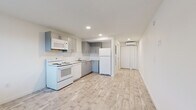 This single-floor unit presents a clean, modern aesthetic defined by a consistent palette of whites and light neutrals throughout. The open kitchen anchors the main living area and is outfitted with gray cabinetry, a white subway tile backsplash, and a full suite of white appliances including an electric range, over-the-range microwave, dishwasher, and refrigerator. Light-toned wood-style flooring runs continuously throughout the space, contributing to an airy, cohesive feel. Recessed lighting illuminates the kitchen and living area, while a window at the front of the unit allows natural light to filter in, offering views of surrounding greenery. A wall-mounted split AC unit provides climate control in the main living zone. The bedroom area is a generous, open room with a closet and additional access to the hallway. The bathroom is finished in all-white tiling, featuring a walk-in shower with large-format tile flooring, a sleek vanity with an undermount sink, and a well-lit mirror. The overall layout flows efficiently from the living and kitchen area through to the private bedroom and bathroom, making the most of the available footprint in a well-maintained, move-in ready condition.