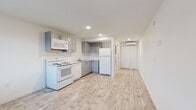 This single-floor unit presents a clean, modern aesthetic defined by a consistent palette of whites and light neutrals throughout. The open kitchen anchors the main living area and is outfitted with gray cabinetry, a white subway tile backsplash, and a full suite of white appliances including an electric range, over-the-range microwave, dishwasher, and refrigerator. Light-toned wood-style flooring runs continuously throughout the space, contributing to an airy, cohesive feel. Recessed lighting illuminates the kitchen and living area, while a window at the front of the unit allows natural light to filter in, offering views of surrounding greenery. A wall-mounted split AC unit provides climate control in the main living zone. The bedroom area is a generous, open room with a closet and additional access to the hallway. The bathroom is finished in all-white tiling, featuring a walk-in shower with large-format tile flooring, a sleek vanity with an undermount sink, and a well-lit mirror. The overall layout flows efficiently from the living and kitchen area through to the private bedroom and bathroom, making the most of the available footprint in a well-maintained, move-in ready condition.