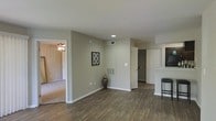 This single-floor residence offers a practical and comfortable layout designed for everyday living. The main living area features rich, dark hardwood-style flooring that extends throughout the open space, creating a cohesive and polished look. Neutral, warm-toned walls provide a versatile backdrop that complements a variety of interior styles.

A recessed ceiling light illuminates the central living area, while a visible HVAC vent and thermostat panel reflect thoughtful climate control integration. A doorway leads into an adjacent room finished with soft carpeting and a ceiling fan, offering a quieter, more comfortable retreat — ideal for use as a bedroom or secondary living space.

Toward one end of the space, a door opens to reveal a walk-in closet with built-in shelving, providing generous storage solutions. A kitchen area is partially visible, featuring a raised breakfast bar with seating, suggesting an open-concept design that keeps the living and dining areas connected. Dark cabinetry accents the kitchen, adding contrast to the lighter walls and countertops.

Wide baseboards and clean wall finishes throughout contribute to the overall neat and well-maintained character of the space. The open flow between rooms and the abundance of natural details make this a functional and inviting single-floor home.