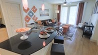 Located at 2010 S. Corinth Street in Corinth, Texas, this single-floor residence offers a thoughtfully designed open-concept layout that seamlessly connects the kitchen and living areas. The kitchen is a standout feature, showcasing a striking curved black granite countertop island that doubles as a breakfast bar, complete with an undermount sink and modern fixtures. The bold, dark surface creates a dramatic contrast against the light-toned walls and flooring, giving the space a contemporary edge.

The living area flows naturally from the kitchen, anchored by a plush sofa, a glass-top coffee table, and a statement zebra-print area rug that adds visual interest to the room. Warm amber and brown accent tones are carried throughout via decorative ottomans, drapery, and wall art, creating a cohesive and vibrant color palette. A ceiling fan with light fixture keeps the space comfortable, while large windows dressed with floor-length curtains invite abundant natural light and provide access to an outdoor area.

Pendant lighting over the island and an arc floor lamp in the living area contribute to a warm, layered lighting scheme. The overall ambiance is bold, modern, and inviting, with carefully curated décor that adds personality and style to every corner of this well-appointed single-level home.