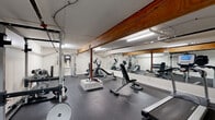 Located at 916 Main St in Buffalo, NY, this fitness facility provides a practical and fully equipped workout environment within a single open floor. The space features durable rubber flooring throughout, bright fluorescent lighting, and white-painted walls that contribute to a clean, functional atmosphere. A prominent wooden beam runs along the ceiling, adding an industrial character consistent with the building's architectural heritage, while exposed structural columns are integrated into the layout.

The gym is outfitted with a comprehensive range of equipment to support diverse training goals. Cardio options include a TRUE treadmill and multiple elliptical machines, while strength training is well-served by a full power rack with Olympic barbell and weight plates, an adjustable bench, and a multi-functional cable machine. A dedicated dumbbell area features a tiered rack stocked with a full set of hex dumbbells. Full-length mirrors line one wall, enhancing the sense of space and allowing users to monitor their form during workouts. A separate doorway leads to an adjacent utility or storage area, adding to the facility's functionality. The overall layout is efficiently organized to maximize usable workout area, making this a well-appointed fitness space suitable for a range of strength and cardio training needs.