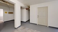 Located at 7594 Watson Rd in Saint Louis, Missouri, this utility room is a single-floor space designed to support the essential mechanical and laundry needs of the building. The room is finished with white-painted walls and a combination of smooth gray-painted concrete and tile flooring, creating a clean and functional environment.

The laundry area is equipped with a commercial-grade washer and dryer, accompanied by a flexible dryer exhaust duct that vents through the wall. A bank of electrical meters is mounted along one wall, providing individual utility monitoring for multiple units within the building. A rolling laundry cart offers added convenience for users.

A dedicated mechanical alcove houses a large commercial water heater with visible copper supply lines and PVC drain piping, reflecting the space's role in supporting building-wide infrastructure. Exposed overhead piping and structural elements run throughout the ceiling, emphasizing the utilitarian character of the space.

Access is provided through solid six-panel doors, and the layout includes open passageways connecting different functional zones within the room. A window with a blind allows for natural light in the laundry area. Overall, the space is well-organized and efficiently designed to serve its operational purpose within the building.