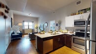 This single-floor residence is thoughtfully designed with an open-concept layout that seamlessly connects the kitchen and living areas. The kitchen is a functional centerpiece, featuring dark wood cabinetry contrasted by crisp white upper cabinets and light countertops. A peninsula with an integrated sink, stainless steel appliances—including a dishwasher, range, microwave, and refrigerator—and bar seating make it well-suited for both cooking and casual dining. Warm hardwood flooring flows throughout the main living space, complementing the neutral gray walls and adding a cohesive warmth to the interior.

The living area is generously proportioned, with tall ceilings fitted with track lighting, a large window allowing natural light to filter in, and ample space for comfortable seating arrangements. The bedroom carries a distinct character, featuring an exposed concrete accent wall, pendant lighting, and a window dressed with layered blinds and sheer curtains that provide a soft, intimate ambiance. The bathroom is clean and contemporary, equipped with a glass-enclosed tiled shower, light tile flooring, dark vanity countertop, and built-in shelving for storage.