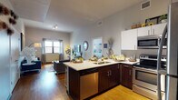 This single-floor residence is thoughtfully designed with an open-concept layout that seamlessly connects the kitchen and living areas. The kitchen is a functional centerpiece, featuring dark wood cabinetry contrasted by crisp white upper cabinets and light countertops. A peninsula with an integrated sink, stainless steel appliances—including a dishwasher, range, microwave, and refrigerator—and bar seating make it well-suited for both cooking and casual dining. Warm hardwood flooring flows throughout the main living space, complementing the neutral gray walls and adding a cohesive warmth to the interior.

The living area is generously proportioned, with tall ceilings fitted with track lighting, a large window allowing natural light to filter in, and ample space for comfortable seating arrangements. The bedroom carries a distinct character, featuring an exposed concrete accent wall, pendant lighting, and a window dressed with layered blinds and sheer curtains that provide a soft, intimate ambiance. The bathroom is clean and contemporary, equipped with a glass-enclosed tiled shower, light tile flooring, dark vanity countertop, and built-in shelving for storage.