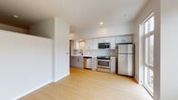 This contemporary single-floor unit offers a clean, modern aesthetic defined by crisp white walls, light wood flooring, and an open-concept layout that maximizes the sense of space. The kitchen is well-appointed with full-height white upper cabinetry, warm wood-toned lower cabinets, a tile backsplash, and a complete suite of stainless steel appliances including a gas range, built-in microwave, dishwasher, and refrigerator. A deep utility sink and recessed lighting complete the functional yet stylish kitchen space. Large floor-to-ceiling windows and additional oversized windows in the living area flood the interior with natural light and provide views of the surrounding neighborhood. The open living space is generous and versatile, with clean lines and neutral tones that offer a blank canvas for any layout. A dedicated hallway connects the main living areas to the private spaces, flanked by full-height built-in storage cabinetry offering ample organizational capacity, as well as a closet with built-in shelving. The bathroom features a wood-toned vanity with a white countertop, a large mirror, and distinctive hexagonal tile flooring in soft gray tones, creating a calm and modern atmosphere. Throughout the unit, recessed lighting, clean baseboards, and consistent finishes reinforce a cohesive, well-designed interior.