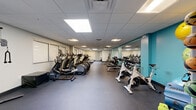 This fitness center at 21200 Brookpark Rd in Cleveland, Ohio offers a comprehensive exercise space designed to support a full range of workout routines. The room features a drop-panel ceiling with recessed fluorescent lighting that provides bright, even illumination throughout. The floors are finished with durable dark rubber gym flooring, well-suited for high-traffic exercise use.

The space is thoughtfully organized into distinct workout zones. The cardio section is generously stocked with treadmills, elliptical cross-trainers, and spinning bikes, offering ample options for cardiovascular training. A strength and free weights area features a weight rack with dumbbells, barbells, a multi-use weight bench, and a cable machine, catering to a variety of resistance training needs. A dedicated functional fitness zone houses medicine balls, stability balls, resistance bands, and additional accessories mounted along a teal accent wall, which adds a pop of color to the otherwise neutral interior palette.

Large mirrors line portions of the walls, enhancing the sense of openness and allowing users to monitor their form. The layout provides generous open floor space between equipment, ensuring comfortable movement throughout the room. The overall ambiance is clean, organized, and purpose-built for fitness, making this a well-rounded facility suitable for a wide range of users and training goals.