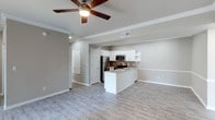This single-story, 2-bedroom, 2-bathroom home features an open-concept design that seamlessly connects the living area and kitchen, creating a spacious and airy feel. Light-toned wood-look flooring runs throughout the main living space, contributing to a clean, contemporary aesthetic.

The kitchen is thoughtfully appointed with white cabinetry, including open display shelving, and is equipped with stainless steel appliances — including a refrigerator, microwave, and range. A tile backsplash adds texture and visual interest behind the cooking area, while light-colored countertops provide ample prep space. A peninsula-style counter extends into the open living area, offering casual seating or additional workspace.

Crown molding lines the ceilings, adding a refined architectural touch throughout the main area. A ceiling fan with a light fixture is centrally positioned, promoting air circulation and supplemental lighting. Neutral gray wall tones create a versatile backdrop suitable for a variety of décor styles.

Doorways visible from the main area lead to additional rooms, including what appears to be a hallway connecting to the bedrooms and bathrooms. The overall layout is efficient and well-organized, making practical use of the available space.