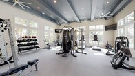 This expansive fitness center is designed to deliver a full-service workout experience in a bright, open environment. The space features soaring ceilings with striking dark-painted exposed beams that contrast elegantly against crisp white walls, creating a dynamic architectural statement. Oversized windows line multiple walls, flooding the interior with natural light and offering views of the surrounding lush landscaping, palm trees, and an adjacent pool area.

The workout floor is covered in durable light grey carpeting and is thoughtfully organized to accommodate a wide range of fitness activities. Cardio equipment includes treadmills, a stationary bike, and a rowing machine, while strength training is well supported with a multi-station cable machine, a Smith machine, selectorized weight machines, TRX suspension trainers, and an Everlast heavy punching bag. A dedicated free weights area features a full rack of dumbbells and medicine balls, paired with adjustable benches.

Large wall-mounted mirrors in the strength zone enhance the sense of space and allow users to monitor their form. Ceiling fans and recessed lighting are distributed throughout, ensuring comfort and visibility. A wall-mounted television provides entertainment during workouts. The overall ambiance is clean, modern, and energizing — a well-appointed facility suited for comprehensive fitness training.