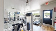 Located at 3331 Luella Blvd in La Porte, Texas, this fitness center offers a thoughtfully designed exercise space across a single floor. The room is finished with light wood-style flooring, crisp white walls accented by a subtle tile wainscoting detail, and crown molding that adds a polished touch to the overall aesthetic. Multiple ceiling fans are distributed throughout, ensuring comfortable airflow during workouts, while recessed lighting provides consistent illumination across the space.

Large windows line the walls, flooding the interior with natural light and offering pleasant views of the surrounding greenery — creating an energizing atmosphere for exercise. The layout is well-organized, with patterned area rugs defining distinct workout zones. The cardio section features multiple treadmills, elliptical machines, and a stationary bike, while the strength training area is equipped with Precor cable machines, a power rack, adjustable benches, a full dumbbell rack, and kettlebells of varying weights. Medicine balls and additional free weights round out the functional training options. A wall-mounted television provides entertainment during workouts. The space strikes a balance between functionality and comfort, making it a comprehensive facility for a wide range of fitness routines.