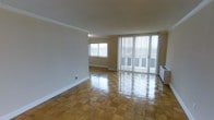 This single-floor space combines classic design elements with an open, airy layout. The main living area is anchored by beautifully polished parquet wood flooring arranged in a traditional block pattern, lending warmth and character to the room. Crown molding runs along the ceiling perimeter, adding a refined architectural detail that elevates the overall aesthetic.

Natural light is a defining feature of this space, with a large window and expansive floor-to-ceiling sliding glass doors fitted with vertical blinds. The sliding doors open to a private balcony, offering outdoor views and allowing abundant light to flood the interior throughout the day.

The neutral wall tones provide a versatile backdrop that complements a wide range of interior styles. The generous proportions of the main living area create a comfortable sense of openness, making it well-suited for both relaxed everyday living and entertaining. A baseboard heating unit is discreetly integrated along the exterior wall, ensuring year-round comfort. Overall, the space presents a clean, well-maintained environment with timeless finishes and a bright, welcoming ambiance.