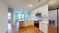 This thoughtfully designed single-floor residence showcases a seamless open-concept layout that connects the kitchen and living area, creating a spacious and functional flow throughout. The kitchen is a standout feature, outfitted with crisp white shaker cabinetry accented by matte black hardware, bright white subway tile backsplash, and clean quartz countertops. A full suite of stainless steel appliances — including a French door refrigerator, electric range, over-the-range microwave, and dishwasher — adds a polished, modern touch. Pendant lighting in warm gold tones hangs above the kitchen peninsula, complementing the light blue accent walls and warm hardwood-style flooring that runs throughout the unit. The open living area is generously proportioned, with natural light streaming in through large windows dressed with blinds. The bedrooms offer comfortable, well-lit spaces, each benefiting from natural light and ceiling fans. The bathroom features a sleek, large-format vanity with a stone countertop, undermount sink, matte black fixtures, and a subway-tiled walk-in shower enclosed by a dark-framed door, delivering a clean and contemporary aesthetic throughout.
