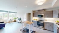 This contemporary single-floor residence showcases a clean, modern design with a bright and open layout that maximizes both comfort and functionality. The open-concept kitchen is well-appointed with wood-grain cabinetry, white quartz countertops, a white tile backsplash, and a full suite of stainless steel appliances including a range, microwave, dishwasher, and refrigerator. A dedicated pantry with built-in shelving provides ample storage, while a compact kitchen island with a butcher-block top serves as a casual dining area. The living and dining spaces flow naturally from the kitchen, benefiting from large windows that frame sweeping views of the surrounding tree-lined neighborhood and rolling hills beyond. The primary bedroom features direct balcony access through a sliding glass door, offering outdoor views, while the second bedroom is carpeted and includes a closet. Both bedrooms are finished in neutral tones with clean lines. The residence includes two bathrooms: a full bath with a glass-enclosed shower, wood-tone vanity, and light tile flooring, and a half bath with a floating vanity and large mirror. Light hardwood flooring runs throughout the main living areas, contributing to the airy, cohesive aesthetic of the space.