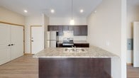Located in East Bethel, Minnesota, this single-floor unit offers a well-designed, open-concept layout that connects the kitchen and living areas seamlessly. The kitchen is equipped with stainless steel appliances, including a refrigerator and range, complemented by rich dark cabinetry that extends to the upper walls for ample storage. A large island with a double sink and granite-style countertops serves as both a functional workspace and a natural divider between the kitchen and the living area. Pendant lighting adds a refined touch overhead. The living area features vaulted ceilings that enhance the sense of openness, with plush carpeting underfoot and large vertical-blind-covered sliding doors that invite natural light. Hardwood-style flooring flows through the kitchen and entry areas, providing a clean contrast to the carpeted spaces. The bedrooms are generously proportioned, carpeted for comfort, and include baseboard heating and natural light from well-placed windows. Two bathrooms serve the unit — one featuring a full tub-shower combo with tile flooring, and another with a walk-in closet access visible through the mirror, white countertops, and dark cabinetry. Warm neutral tones and consistent finishes throughout lend the space a cohesive, move-in-ready aesthetic.