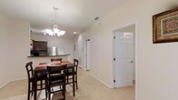 This single-floor residence offers a clean, functional layout with a warm and neutral aesthetic throughout. The open-concept dining and kitchen area serves as the heart of the home, anchored by a elegant chandelier with frosted glass shades that provides warm overhead lighting above a dark wood dining table with seating for four. Light-toned ceramic tile flooring extends across the main living areas, lending a bright and cohesive feel to the space.

The kitchen features dark cabinetry with a contrasting countertop, a stainless steel refrigerator, and a tile backsplash, offering a practical and stylish cooking environment. A breakfast bar connects the kitchen seamlessly to the dining area, making it ideal for casual meals or entertaining.

A central hallway extends from the main living area, providing access to the additional rooms within the home. Crisp white six-panel doors and white baseboards throughout contribute to a clean, well-maintained appearance. The walls are finished in soft, neutral tones that enhance the sense of openness and allow natural and recessed lighting to brighten the interior effectively. Ceiling vents indicate a central HVAC system, ensuring year-round comfort. The overall layout is practical and inviting, suitable for everyday living.