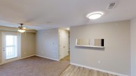 This single-floor space is designed with a clean, functional layout that emphasizes an open and connected feel. The living area is finished with plush carpeting in a neutral tone, while the adjacent entryway and transitional spaces feature smooth hardwood-style flooring, creating a clear visual distinction between zones. The walls are painted in a warm greige tone throughout, contributing to a cohesive and calming atmosphere.

A notable architectural detail is the pass-through window fitted with a granite-style ledge, which connects the living area to the kitchen, promoting an open flow between spaces while maintaining defined areas. Recessed lighting and a flush-mount ceiling fixture provide consistent illumination across the space.

The entryway leads to what appears to be a closet with classic paneled doors, offering practical storage upon entry. A floor vent is positioned along the baseboard, indicating a forced-air climate control system. White baseboards and trim provide clean contrast against the neutral walls, adding a polished, finished look throughout. Overall, the space presents a straightforward and well-maintained interior suited for comfortable everyday living.