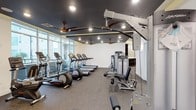 This fitness center is thoughtfully designed to support a well-rounded workout experience. The space is anchored by a row of cardio machines, including treadmills, ellipticals, and a stationary bike, all positioned along a wall of large floor-to-ceiling windows that flood the room with natural light and offer views of the surrounding exterior. A dedicated strength training area rounds out the equipment selection, featuring a multi-function cable machine and additional resistance equipment, providing users with comprehensive options for both upper and lower body training. A storage cubby unit is also available for convenient organization of personal belongings. The interior design strikes a bold contrast, with dark-painted ceilings fitted with recessed lighting and multiple ceiling fans that keep the environment comfortable during intense workouts. The flooring consists of durable rubber gym matting, offering both safety and functionality underfoot. The overall ambiance is modern and purposeful, combining practical amenities with a clean, motivating atmosphere well-suited for daily fitness use.