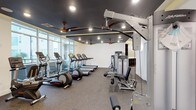 This fitness center is thoughtfully designed to support a well-rounded workout experience. The space is anchored by a row of cardio machines, including treadmills, ellipticals, and a stationary bike, all positioned along a wall of large floor-to-ceiling windows that flood the room with natural light and offer views of the surrounding exterior. A dedicated strength training area rounds out the equipment selection, featuring a multi-function cable machine and additional resistance equipment, providing users with comprehensive options for both upper and lower body training. A storage cubby unit is also available for convenient organization of personal belongings. The interior design strikes a bold contrast, with dark-painted ceilings fitted with recessed lighting and multiple ceiling fans that keep the environment comfortable during intense workouts. The flooring consists of durable rubber gym matting, offering both safety and functionality underfoot. The overall ambiance is modern and purposeful, combining practical amenities with a clean, motivating atmosphere well-suited for daily fitness use.