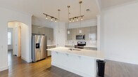 This two-story space showcases a contemporary design aesthetic defined by clean lines, neutral tones, and quality finishes throughout. The kitchen serves as a focal point, featuring a large central island with a crisp white quartz countertop and an integrated sink. Gray flat-front cabinetry extends to the upper walls, providing generous storage, while a marble-look tile backsplash adds visual interest. Stainless steel appliances, including a French door refrigerator with water dispenser, an electric range, and a built-in microwave, complete the well-appointed cooking space. Brass-finish pendant lights and track lighting illuminate the kitchen, adding a warm contrast to the cool-toned cabinetry. Wood-look flooring flows consistently through the main areas, grounding the light color palette. The bathrooms maintain the modern theme — one features a pedestal sink, a mirror with vanity lighting, and a window providing natural light, while a second bathroom includes grab bars and a floating vanity. Additional rooms present bright, open spaces with white walls, dark hardwood-style floors, and windows fitted with horizontal blinds. The overall ambiance is clean, minimal, and move-in ready.