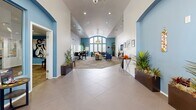 Located at 1155 W Grove Pkwy in Tempe, Arizona, this single-floor community clubhouse offers a thoughtfully designed gathering space that combines comfort with a polished aesthetic. The layout flows seamlessly from a tiled entry foyer into a generously sized open lounge area, where soaring vaulted ceilings and large arched windows flood the interior with natural light, creating an airy and inviting atmosphere.

The lounge is anchored by a patterned area rug and furnished with plush seating including a neutral sofa, navy blue accent chairs, and a round coffee table, all centered beneath a stylish drum pendant light. A dining area with bar-height tables and chairs is positioned adjacent to the lounge, ideal for casual gatherings. The accent walls painted in a rich blue-gray tone provide a striking contrast to the crisp white walls and ceilings throughout.

A reception counter with an arched pass-through serves as a functional hub, complemented by a water station and a built-in coffee setup for convenience. A wall-mounted television and framed artwork add character to the space. The office, accessible via arched doorways, provides a quieter workspace, while the kitchen supports the overall functionality of the clubhouse. Potted tropical plants throughout the space add warmth and a touch of vibrant greenery.