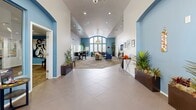 Located at 1155 W Grove Pkwy in Tempe, Arizona, this single-floor community clubhouse offers a thoughtfully designed gathering space that combines comfort with a polished aesthetic. The layout flows seamlessly from a tiled entry foyer into a generously sized open lounge area, where soaring vaulted ceilings and large arched windows flood the interior with natural light, creating an airy and inviting atmosphere.

The lounge is anchored by a patterned area rug and furnished with plush seating including a neutral sofa, navy blue accent chairs, and a round coffee table, all centered beneath a stylish drum pendant light. A dining area with bar-height tables and chairs is positioned adjacent to the lounge, ideal for casual gatherings. The accent walls painted in a rich blue-gray tone provide a striking contrast to the crisp white walls and ceilings throughout.

A reception counter with an arched pass-through serves as a functional hub, complemented by a water station and a built-in coffee setup for convenience. A wall-mounted television and framed artwork add character to the space. The office, accessible via arched doorways, provides a quieter workspace, while the kitchen supports the overall functionality of the clubhouse. Potted tropical plants throughout the space add warmth and a touch of vibrant greenery.