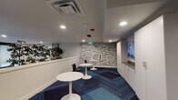 Located at 400 S Jackson St in Seattle's historic Pioneer Square neighborhood, this single-floor amenity space is thoughtfully designed to serve residents with a range of practical and social conveniences. Upon entry, a bank of smart package lockers provides secure, self-service parcel retrieval, positioned alongside elevator access for ease of use.

The lounge area features a vibrant kitchenette with a bold yellow-green countertop, ample cabinetry, a stainless steel appliance, and a sink — accented by a trio of colorful abstract artworks mounted above. A peninsula counter with bar seating encourages casual interaction. The patterned carpet throughout incorporates a dynamic mix of blue, teal, and charcoal hues in a chevron-inspired layout that adds visual energy to the space.

A dedicated lounge nook offers round pedestal tables and mixed seating, anchored by a striking typographic accent wall featuring Seattle-inspired words and phrases — a nod to the city's iconic culture and landmarks. Lush indoor trees line a row of large windows, framing views of the surrounding cityscape while bringing a refreshing natural element indoors. Wall-mounted flat-screen displays are positioned throughout for informational or entertainment use. Recessed lighting throughout ensures a bright, welcoming atmosphere across the entire space.