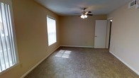 This single-floor residential space offers a straightforward and functional layout designed for comfortable everyday living. The main living area is finished with wall-to-wall carpeting in a neutral tone, providing a warm and cohesive foundation throughout the room. Smooth, light-colored walls in a soft beige hue lend a clean, airy feel to the interior, while white baseboards add a crisp finishing touch along the perimeter. A large window allows generous natural light to flow into the space, casting bright patches across the floor and enhancing the open, welcoming ambiance of the room. A ceiling fan with an integrated light fixture is centrally mounted, offering both practical ventilation and supplemental lighting. A white interior door is visible along one wall, suggesting additional rooms or storage beyond the main living area. The overall atmosphere is clean, modest, and move-in ready, making it a versatile space well-suited for a variety of living arrangements. The simple, uncluttered design allows for flexible furniture placement and personalization to suit individual tastes.