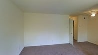 This single-floor residential space offers a clean, understated interior defined by neutral white walls and wall-to-wall carpeting throughout. The image reveals a spacious, open room with a simple and functional design aesthetic. The smooth white walls provide a bright, airy atmosphere, offering a versatile blank canvas that adapts well to a variety of furnishing styles and décor preferences. The room benefits from generous proportions, giving a comfortable sense of openness and flexibility in how the space can be used. A doorway visible along one wall leads to an additional area, which appears to include a closet or storage space, adding practical utility to the layout. The neutral carpet underfoot contributes warmth and a quiet, residential feel throughout. The space is characterized by its straightforward, unfussy design — clean lines, minimal architectural detailing, and a cohesive color palette of whites and soft grays that work together to create a calm, livable environment. The overall ambiance is bright and welcoming, well-suited for everyday residential use.