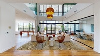 The Natura Gardens Clubhouse in Hialeah, Florida is a thoughtfully designed single-story community facility that blends modern architecture with a warm, resort-inspired aesthetic. The entrance greets visitors with warm hardwood flooring, curated wall art, and lush tropical greenery framing glass-paneled doors that frame views of the surrounding landscape.

The main lounge is the centerpiece of the clubhouse, boasting dramatic double-height ceilings and expansive floor-to-ceiling windows that flood the space with natural light and offer views of the palm-lined outdoors. Oversized woven pendant lights hang above a seating arrangement of plush neutral sofas, rattan accent chairs, and a bold zebra-print area rug, creating an inviting yet vibrant atmosphere. A billiard table adds a recreational element to the space.

A glass-enclosed co-working area provides a dedicated workspace with a conference-style table and ergonomic seating, maintaining visual connectivity with the main lounge. Adjacent to this is an additional lounge area featuring comfortable sectional seating and curated artwork.

The clubhouse kitchen is clean and contemporary, featuring white cabinetry, stainless steel appliances, a spacious center island with bar seating, and sleek black pendant lighting—ideal for community gatherings and events.