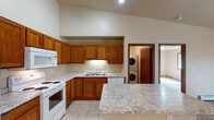 This single-floor residence offers a functional and comfortable layout designed for everyday living. The heart of the home is an open-concept kitchen and living area with soaring vaulted ceilings that create an airy, expansive feel. The kitchen is equipped with warm oak cabinetry, light granite-style countertops, a central island for additional prep space, and a full suite of white appliances including a refrigerator, electric range, built-in microwave, and dishwasher. Light ceramic tile flooring runs throughout the kitchen and utility areas, while soft carpet covers the living space and bedrooms. The living area features a ceiling fan, track lighting, a decorative pendant light, and a baseboard heating system. A dedicated laundry room off the kitchen houses a stacked front-loading washer and dryer alongside a water heater. The primary bathroom offers a spacious vanity with dual sinks, a separate glass-enclosed shower, and direct access to a generously sized walk-in closet with built-in shelving and hanging rods. A second full bathroom includes a tub-shower combo and a single vanity. Bedrooms feature carpeted floors, baseboard heat, and windows that bring in natural light.