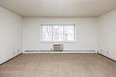 This single-floor space offers a clean, functional layout designed for comfortable everyday living. The main living area is generously sized, finished with neutral wall-to-wall carpeting and crisp white walls beneath a textured ceiling. A large sliding window floods the room with natural light and provides views of mature trees and the surrounding neighborhood, while a baseboard heating system and wall-mounted air conditioning unit ensure year-round climate comfort.

The bedroom is a well-proportioned room featuring soft carpeting, a ceiling fan with light fixture, and a bifold closet with built-in shelving, offering practical storage solutions. A door with a mirrored inset adds a functional and stylish touch.

The galley-style kitchen is efficiently arranged, featuring warm honey-toned wood cabinetry, light countertops, a stainless steel sink, a gas range, and a full-size refrigerator. Tile flooring runs throughout the kitchen, lending durability and easy maintenance.

The bathroom is neatly finished with tile flooring, a warm wood vanity with a light stone-topped counter, a mirrored medicine cabinet, and a chrome wall-mounted shelving rack that provides additional storage. The overall aesthetic throughout the space is neutral and understated, offering a versatile canvas ready to be personalized.