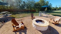This community amenity space combines an indoor fitness center with an inviting outdoor fire pit area, offering residents a well-rounded recreational experience. The fitness center features a bold color scheme with striking orange and blue accent bands along the upper walls, complemented by neutral tones and patterned carpet flooring throughout. Large windows along one wall flood the space with natural light and provide views of the outdoor pool area, creating an energizing workout environment. The room is equipped with a comprehensive selection of cardio machines, including multiple treadmills, upright and recumbent stationary bikes, an elliptical trainer, and a rowing machine. Strength training is well represented with a multi-station cable machine, a weight bench, and a full rack of dumbbells displayed in front of a large wall-mounted mirror, which enhances the sense of openness. A ceiling fan and recessed lighting ensure comfort during workouts. The space also includes a fire safety station near the entrance door. Outside, the fire pit area centers around a substantial circular stone-built fire pit surrounded by natural gravel ground cover. Wooden Adirondack chairs are arranged around the pit for relaxed gatherings, while a shaded picnic table with a blue umbrella and additional seating are situated nearby. Mature trees and a wooden privacy fence enclose the outdoor space, creating a secluded and comfortable setting for social gatherings.