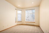 This single-floor residential unit offers a practical and well-organized layout designed for comfortable everyday living. A carpeted hallway with recessed lighting and warm wood-toned sliding storage doors leads into the main living areas, setting a clean and inviting tone from the moment you enter.

The open-concept living space is generously proportioned, featuring plush wall-to-wall carpeting and soaring double-height ceilings that lend an airy, expansive feel. Natural light filters in through large windows, creating a bright and welcoming atmosphere. The kitchen flows seamlessly from the living area via a peninsula-style counter, and is equipped with light wood cabinetry, a coil-top electric range, an over-the-range microwave, a refrigerator, and durable laminate countertops — all set atop easy-care hardwood flooring.

The bedroom is a comfortable, carpeted retreat with a large sliding glass door that opens onto a private balcony, offering views of the surrounding urban streetscape. An additional room features a generous window with a baseboard heater, providing both natural light and practical warmth. The bathroom is clean and functional, appointed with a full tub-shower combination, tile flooring, and a curved shower rod. Throughout the unit, warm wood baseboards and neutral walls create a cohesive, understated aesthetic.