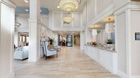 Located at 115 Healthy Way in Folsom, California, this expansive single-floor lobby offers a refined and welcoming atmosphere across a generously sized footprint. The architectural design is defined by soaring double-height ceilings, crisp white columns, and wide-plank light wood-tone tile flooring that flows seamlessly throughout the space.

Upon entry through ornate wood-and-glass doors, visitors are greeted by a grand foyer anchored by a series of oversized pendant lantern chandeliers with gold brass accents. A reception desk finished in decorative granite sits to one side, adorned with fresh floral arrangements. Elegant accent walls featuring botanical murals add a touch of artistry to the entryway seating area, which is furnished with sophisticated wingback chairs.

The space opens into multiple distinct lounge areas, each thoughtfully curated with plush sofas, armchairs in soft blue and blush tones, and patterned area rugs. Tall clerestory windows and a coffered ceiling detail with soft blue accents draw in abundant natural light throughout.

The dining room is a standout feature, lined with floor-to-ceiling wood-framed glass doors that offer direct views of an outdoor pool and palm tree-lined courtyard. Inside, marble-topped tables, striped banquette seating, and blue upholstered chairs create an inviting and sophisticated dining environment bathed in natural light.