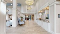 Located at 115 Healthy Way in Folsom, California, this expansive single-floor lobby offers a refined and welcoming atmosphere across a generously sized footprint. The architectural design is defined by soaring double-height ceilings, crisp white columns, and wide-plank light wood-tone tile flooring that flows seamlessly throughout the space.

Upon entry through ornate wood-and-glass doors, visitors are greeted by a grand foyer anchored by a series of oversized pendant lantern chandeliers with gold brass accents. A reception desk finished in decorative granite sits to one side, adorned with fresh floral arrangements. Elegant accent walls featuring botanical murals add a touch of artistry to the entryway seating area, which is furnished with sophisticated wingback chairs.

The space opens into multiple distinct lounge areas, each thoughtfully curated with plush sofas, armchairs in soft blue and blush tones, and patterned area rugs. Tall clerestory windows and a coffered ceiling detail with soft blue accents draw in abundant natural light throughout.

The dining room is a standout feature, lined with floor-to-ceiling wood-framed glass doors that offer direct views of an outdoor pool and palm tree-lined courtyard. Inside, marble-topped tables, striped banquette seating, and blue upholstered chairs create an inviting and sophisticated dining environment bathed in natural light.