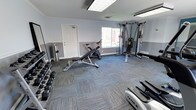 This fitness center offers a comprehensive workout environment designed to support a wide range of exercise routines. The space is thoughtfully organized across a single floor, with a clean, modern aesthetic defined by two-tone gray walls, white crown molding, and multi-toned blue-gray carpet tile flooring throughout.

The cardio zone is well-appointed with True-brand treadmills, an elliptical trainer, and a recumbent bike, positioned near large windows that invite natural light and offer views of the surrounding exterior. A dedicated strength training area features a cable crossover multi-station machine, an adjustable workout bench, and a spinning bike, providing options for both resistance and cycling workouts.

The free weights section includes a full rack of dumbbells in graduated sizes, accompanied by a large floor-length mirror that enhances the sense of space and supports proper form during exercise. Additional functional fitness equipment includes stability balls, medicine balls, exercise mats, and TRX suspension training gear, supporting versatile training modalities.

Overhead fluorescent lighting supplements the natural light from multiple windows, ensuring the space remains well-lit throughout. The facility is finished with a teal accent wall and mirrored wall panel in the cardio area, adding a dynamic visual element while further amplifying the openness of the room.