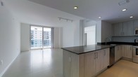 This single-floor space showcases a contemporary open-plan design that seamlessly integrates the kitchen and living areas. The kitchen features a prominent island with a dark countertop and light wood cabinetry, providing ample storage and workspace. A modern faucet adds a sleek finishing touch. The living area is bathed in natural light, courtesy of expansive floor-to-ceiling windows that frame sweeping city views of surrounding buildings. The clean white walls and large-format light-toned tile flooring throughout create a bright, airy ambiance that enhances the sense of space. Recessed lighting and a track lighting fixture provide adjustable illumination, complementing the abundant daylight. The open layout encourages flexible use of the space, with a clear sightline from the kitchen to the living area. A separate room is visible in the background, accessible through a defined opening. The overall design aesthetic is minimalist and modern, with a neutral color palette that highlights the architectural details and keeps the focus on the generous proportions and urban vistas.