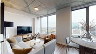 This stylish high-rise residence blends industrial and contemporary design elements across a well-appointed single-floor layout. The living area is a standout, anchored by dramatic floor-to-ceiling windows that frame sweeping panoramic city views, flooding the space with natural light. Exposed concrete ceilings add a distinctive industrial character that contrasts beautifully with the clean white walls and warm wood-tone flooring found throughout.

The open-concept layout connects the living room seamlessly to the dining area and kitchen. The kitchen is outfitted with dark navy cabinetry, light quartz countertops, a tile backsplash, and stainless steel appliances including a smooth-top range and over-the-range microwave. The dining area accommodates a round table with seating for multiple guests.

The bedroom continues the exposed concrete ceiling aesthetic, offering a cozy and intimate atmosphere accented by warm lighting and ample space for a full furniture arrangement. The bathroom features dark large-format floor tiles, a wood-toned vanity with white countertop, an undermount sink, and a large mirror, delivering a clean and modern finish.

Throughout the unit, consistent use of warm wood tones, neutral palettes, and natural textures creates a cohesive, contemporary ambiance well-suited to urban living at elevation.